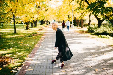 Sarı yapraklı ağaçlarla yol kenarında gülmekten uzun siyah ve çizgili pantolon giymiş genç bir sarışın. Kız gülünç bir poz veriyor ve sonbahar akçaağaçlarının arka planında eğleniyor.