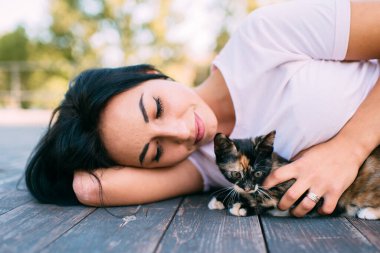Güzel, genç, beyaz bir kız ahşap zeminde küçük bir kedi yavrusuyla yatıyor ve o anın keyfini çıkarıyor.