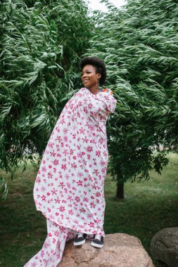 Güzel, koyu tenli, çarşafa sarılmış, arkasında ağaçlarla parkta poz veren bir kız..