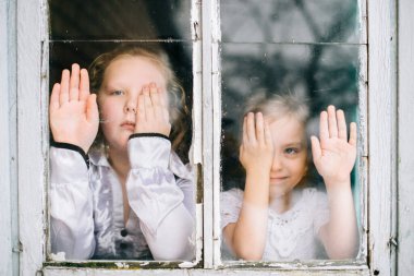 İki küçük kız elleriyle gözlerini kapatıyor ve komik yüz ifadeleriyle pencereye bakıyor. 