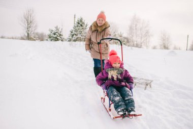 Anne, kızıyla dışarıda karlı bir kış gününde kızağın üzerinde oturmuş eğleniyor..