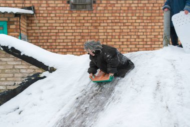 Yetişkin cüssesi ceketli komik çocuk kış günü buzlu yoldan bodruma doğru kızakla iniyordu. Duygusal yüzlü küçük kız köy bahçesinde kayıyor.