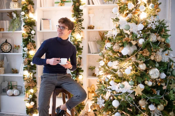 Elegant man with positive emotional face posing for camera with cup of coffee in cozy room with cristmas tree and new year decorations