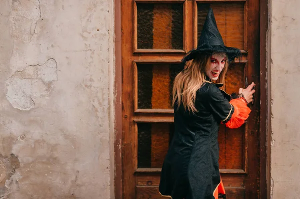 Emotionally witch with big black hat, long brown hair, beautiful eyes and bright makeup poses outside in front of the big old door in black and red dress