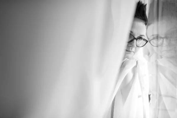 Black and white picture of lovely young caucasian female with pretty face, short dark hair, big eyes lies on the big white sofa wrapped herself in curtains