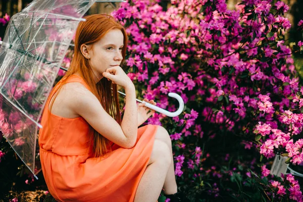 長い赤い髪をした奇妙な若い女の子 夏のオレンジのドレスでかなり顔は庭で傘を保持しています — ストック写真