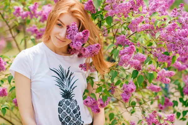 赤い髪とかなり笑顔で美しい若い白人の女の子は 花とライラックの庭にポーズをとって それの匂いを楽しむ — ストック写真