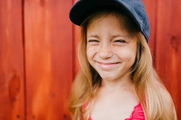 Picture of cute caucasian girl with long fair hair and in pink dress and denim cap smiles and makes faces against the wooden wall in village