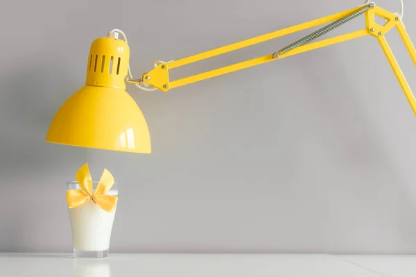 Glass of milk decorated with yellow bowtie standing on table under yellow lamp.
