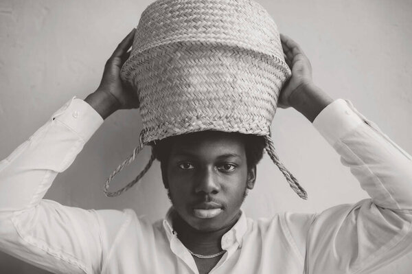Closeup lifestyle portrait of strange young african black man holding straw basket above his head on textured wall background and looking at camera. Dark-skinned boy in white t-shirt funny portrait.