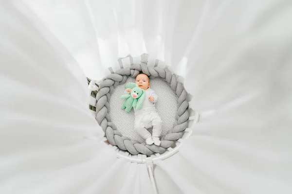 Portrait from above of beautiful funny newborn baby boy lying on back in white round bed with canopy.  Nursery for infant. Kids sleep wear.  Charming cheerful child make funny faces in the morning