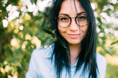 Büyük kahverengi gözlü, uzun koyu renk saçlı, kısa mavi elbiseli, yeşil ağacın yanında siyah gözlüklü, harika bir gülümsemesi olan hoş, genç, beyaz bir kız.