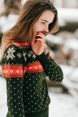 Uzun saçlı, uzun saçlı, desenli Noel süveteri giyen ve karlı kış ormanlarında kameraya poz veren genç bir kız..