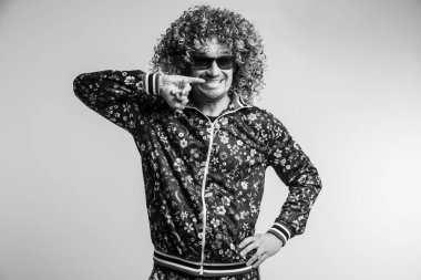 80 'lerin moda eşofmanlı komik bir adamın kapalı portresi çiçek desenli ve güneş gözlüklü parmaklarıyla dişlere dokunan. Yetişkin, neşeli, kıvırcık saçlı, beyaz stüdyo geçmişiyle dalga geçen bir erkek.