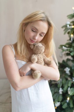 Gecelikli genç kız, arka planda Noel ağacı olan oyuncak ayıya sarılıyor..
