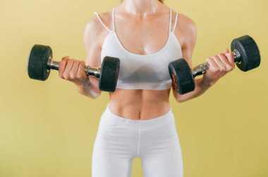 Dambılı atletik vücut geliştiren kadın. Güzel esmer ve kaslı kız sarı arka planda ağırlık kaldırıyor.