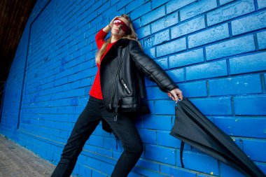 Modayı takip eden esmer bir kızın moda portresi. Koyun derisi kazı ve elinde şemsiyeyle dışarıda mavi tuğla duvara poz veren kırmızı güneş gözlüğü..