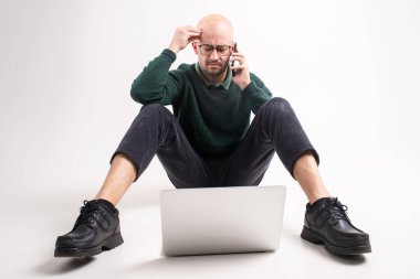 Yakışıklı beyaz bir erkek bir elinde telefonu, diğer elinde laptopuyla tableti, bir katta oturuyor ve beyaz arka planda izole edilmiş telefonu arıyor.