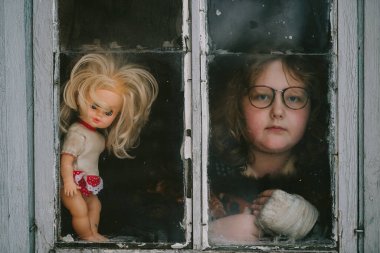 Kırık kollu yalnız bir çocuğun portresi. Eski plastik bir bebekle pencereden dışarı bakıyor.