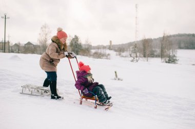 Anne, kızıyla dışarıda karlı bir kış gününde kızağın üzerinde oturmuş eğleniyor..