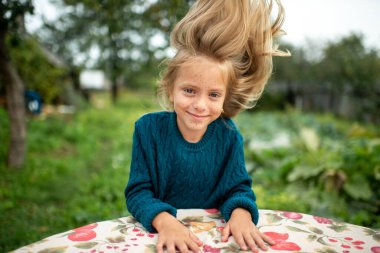 Mavi kapüşonlu, uzun sarı saçlı güzel beyaz kız ağacın yanındaki çarşafta uzanıyor ve saçlarıyla oynuyor.