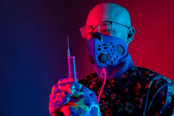 Stylish bald man wearing respirator mask and glasses and holding syringe with vaccine against covid 19 in his hand over red blue background. Coronavirus concept