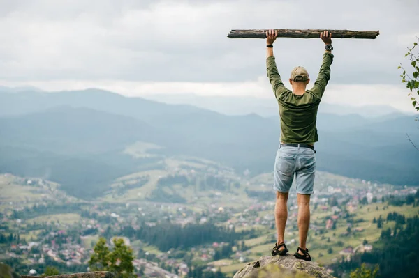 Lifestyle summer portrait from behind of successful man with wooden stick standing on top of mountaing with beautiful landscape in front. Male traveler enjoying nature view from highest peak at hill