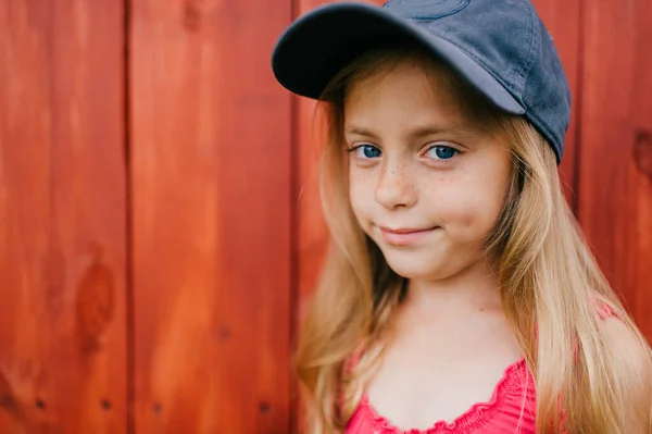 Picture of cute caucasian girl with long fair hair and in pink dress and denim cap smiles and makes faces against the wooden wall in village