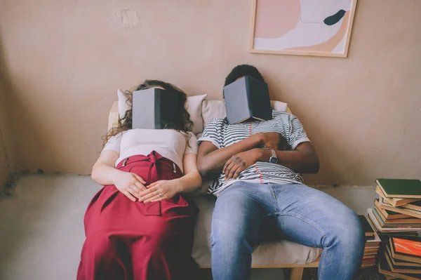 Funny interracial couple sitting in chair in studio and sleeping with books on their faces. Tired dark skinned unrecognizable man fall asleep after reading with caucasian unknown woman at home.
