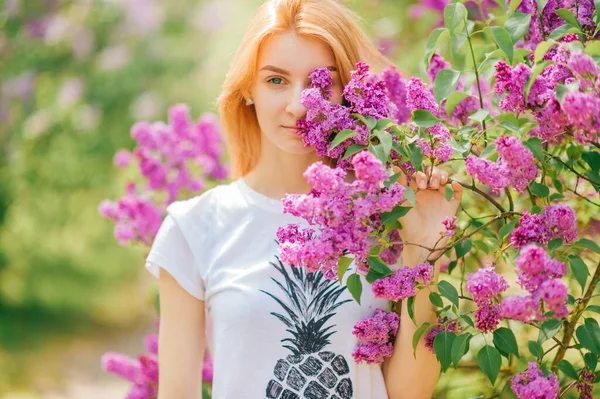 赤い髪とかなり笑顔で美しい若い白人の女の子は 花とライラックの庭にポーズをとって それの匂いを楽しむ — ストック写真