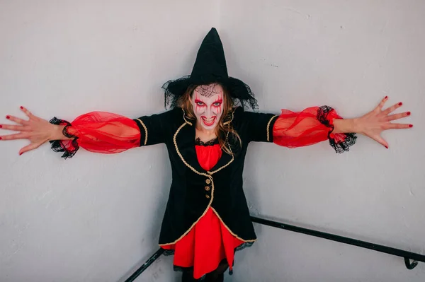 Portrait of a mysterious girl with fair hair as a witch with bright make-up and leaking lipstick mysteriously looks into the camera with wide smile and stands in the corner