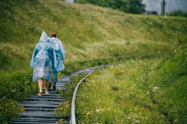 两个穿着雨衣的年轻旅行者走在长满青草的铁路上 一群朋友在度假时休息 旅游服装 多云的闷热天气 希波斯特的生活方式从角度来看哭 — 图库照片