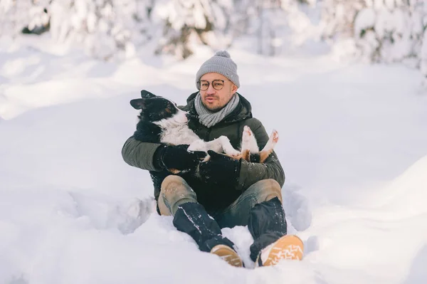Happy man holding lovely dog in his hands in snowy forest. Smiling boy hugging adorable puppy in winter wood. Pet lover.  Dog - human`s friend concept.