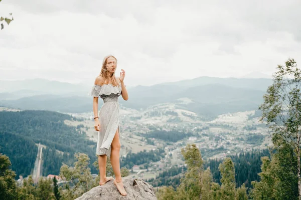 石で征服された山の上に立っている夏のドレスに長い髪を持つ若い美しい裸足のブロンドの女の子と渓谷の山や村で素晴らしい景色を楽しむ — ストック写真