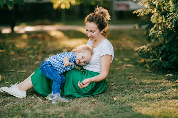 Beautiful caucasian mum with dark hair in white t-shirt and long green skirt plays in the park with her lovely son with short fair hair in white and blue shirt and blue jeans.