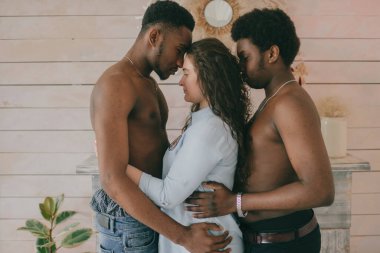 Çok kültürlü aşk ve ilişkiler kavramı. Beyaz seksi kız iki üstsüz koyu tenli adamı kucaklıyor. Irklar arası aşk üçlüsünün kapalı bir portresi. İki tutkulu Afrikalı adam sevgililerine sarılıyor.