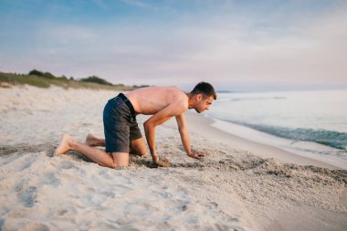 Genç, çıplak ayaklı, şortlu, dizlerinin üzerinde duran ve yaz akşamları denizin ötesindeki kumsalda el pençe divan kazan adam. Okyanus çocuğu gibi ıssız bir yerde kumla sahilde oynayan komik çocuk.. 