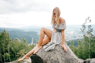 Genç, güzel, uzun saçlı, dağda taşın üzerinde oturan güzel, bulutlu gökyüzü manzaralı sarışın. Yazlık elbiseler içinde hoş bir kadın. Rahatlatıcı ve açık hava portresi. Doğada mutlu bir kadın.