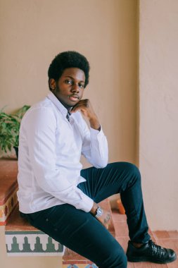 Duvarda desenli dekoratif desenlerle merdivenlerde oturan Afro-Amerikan siyahi bir adamın yaşam tarzı. Mutlu koyu tenli Nijeryalı adamın kapalı oda portresi..