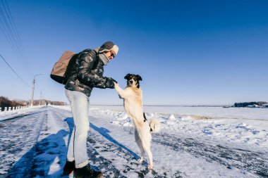 Man in down padded coat, fur cap, blue jeans and sunnglasses dancing with homeless dog on road beside frozen lake in winter snowy sunny day. Cold weather. Funny domestic animal. Pet treatment and care clipart