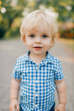 Mavi tişörtlü, mavi şortlu, neşeli beyaz çocuk parkta yürüyüşe çıkıyor..