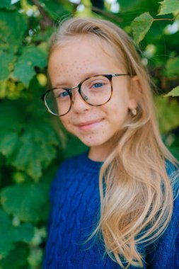Beyaz saçlı, uzun sarı saçlı, mavi kapüşonlu, gözlüklü genç bir kızın portresi yeşil yaprakların arkasındaki bahçede duruyor.