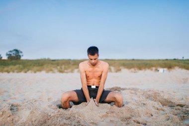 Genç üstsüz bir adam denizin ötesinde kumsalda oturuyor. Bacakları kumun içinde ve elleri çocuk gibi kum heykelleri yapıyor. Hobi, yaz tatili konsepti. Sanat şahsiyeti yaratıcı etkinlik