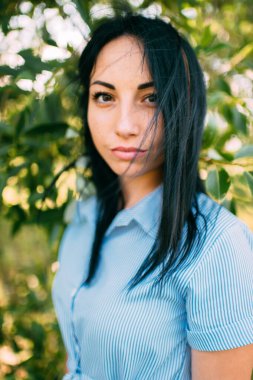 Büyük kahverengi gözlü, uzun siyah saçlı, yakışıklı beyaz bir kızın portresi ve bahçedeki yeşil ağacın yanındaki kısa mavi elbiseyle mükemmel bir gülümseme.