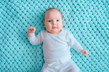 Adorable lovely newborn male baby with blue eyes lifestyle  indoor portrait from above. Cute child with plump cheeks lying on back in white pijama on turqoise woolen plaid. Funny nursing kid emotions. clipart