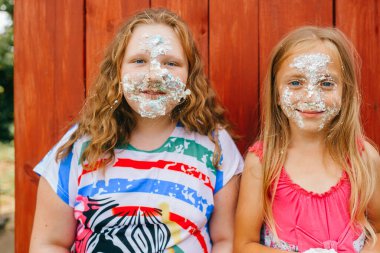Beyaz saçlı, uzun boylu, pembe elbiseli, şişman ablasıyla oynayan ve köyün ahşap duvarına karşı birbirlerinin yüzlerine pasta süren beyaz bir kız resmi.