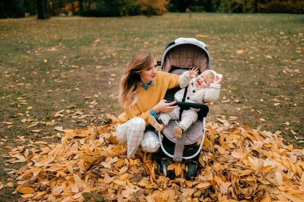 Cute little baby with big blue eyes, short red hair, pump lips in a stroller outside in the fall with nice happy mum sits near it in leaves