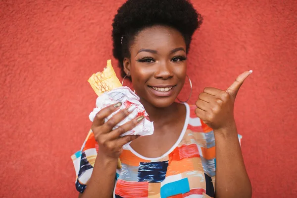 Neşeli Afro Amerikalı Kız Elinde Hamburgerle Arka Plandaki Kırmızı Duvarın — Stok fotoğraf