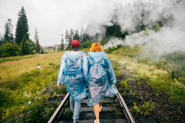 Company of friends walking along railway track. Traveling couple in blue raincoats walk along railroad outdoor at nature in cloudy rainy day. Portrait from behind of man and woman smoking vape.