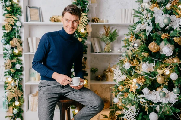 Elegant man with positive emotional face posing for camera with cup of coffee in cozy room with cristmas tree and new year decorations